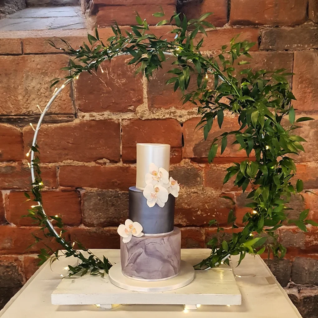 A decorative multi-tiered cake with white and gray marbled tiers, adorned with white orchids, set on a white board on a table with a brick wall background. Green leaves and small string lights decorate the arrangement.