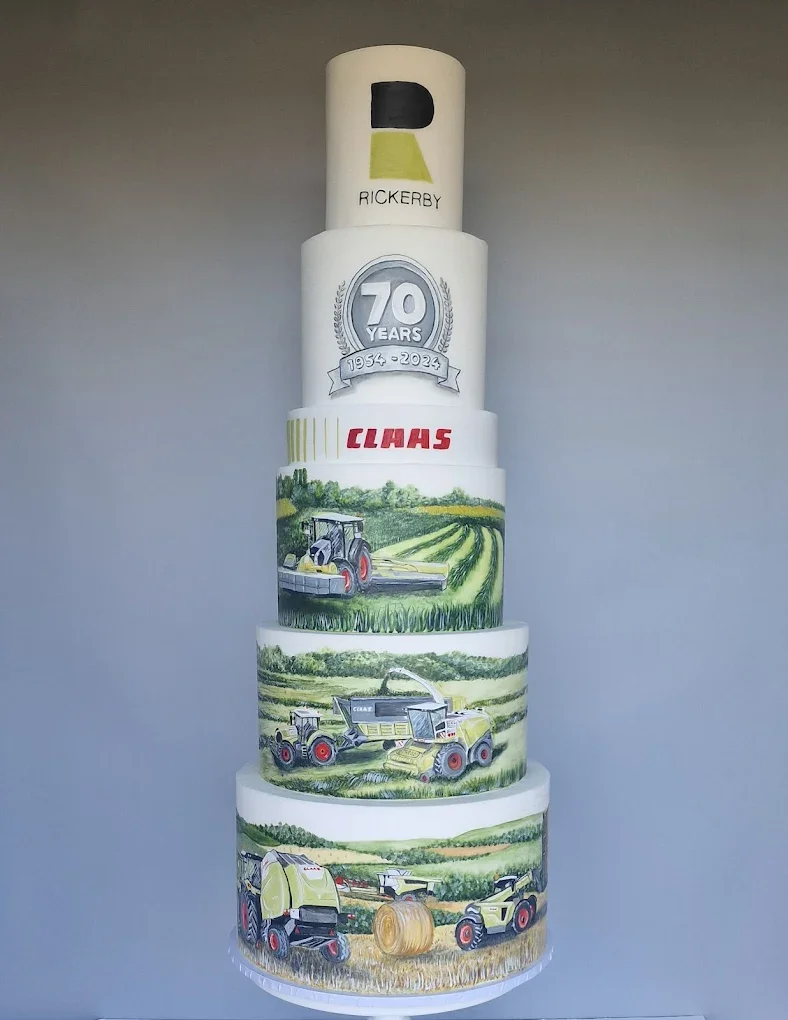 A four-tiered cake celebrating the 70th anniversary of Rickerby, featuring logos and illustrations of agricultural equipment like tractors, a baler, and a sprayer, with a farm landscape painted on the cake layers.