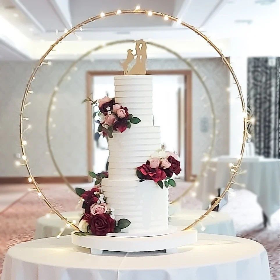 A wedding cake with a topper of a couple kissing, surrounded by pink and red flowers, with two golden rings and small lights in a circular arrangement behind it, on a white table.