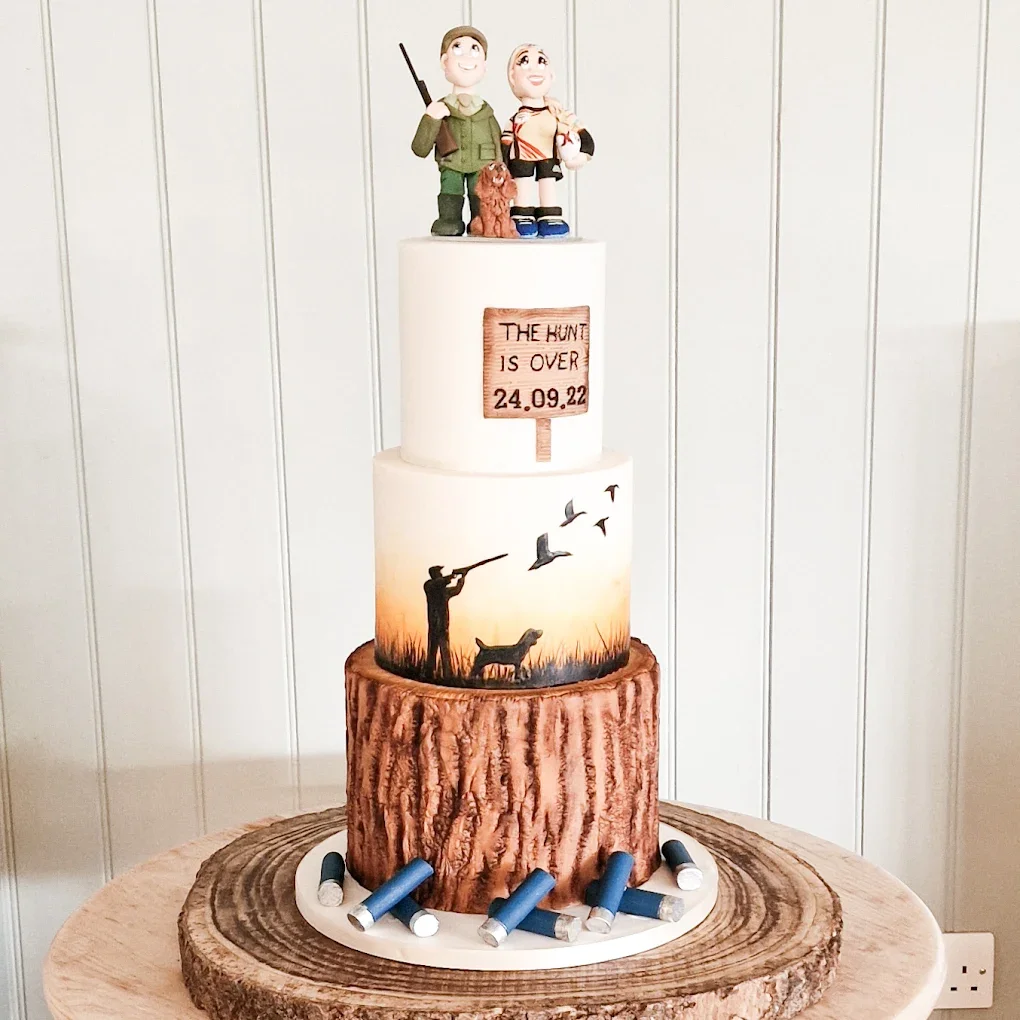 A multi-tiered cake with a woodland hunting theme, featuring figurines of hunters and a dog on top, painted with silhouettes of a hunter and birds, and decorated with fake shotgun shells around the base.