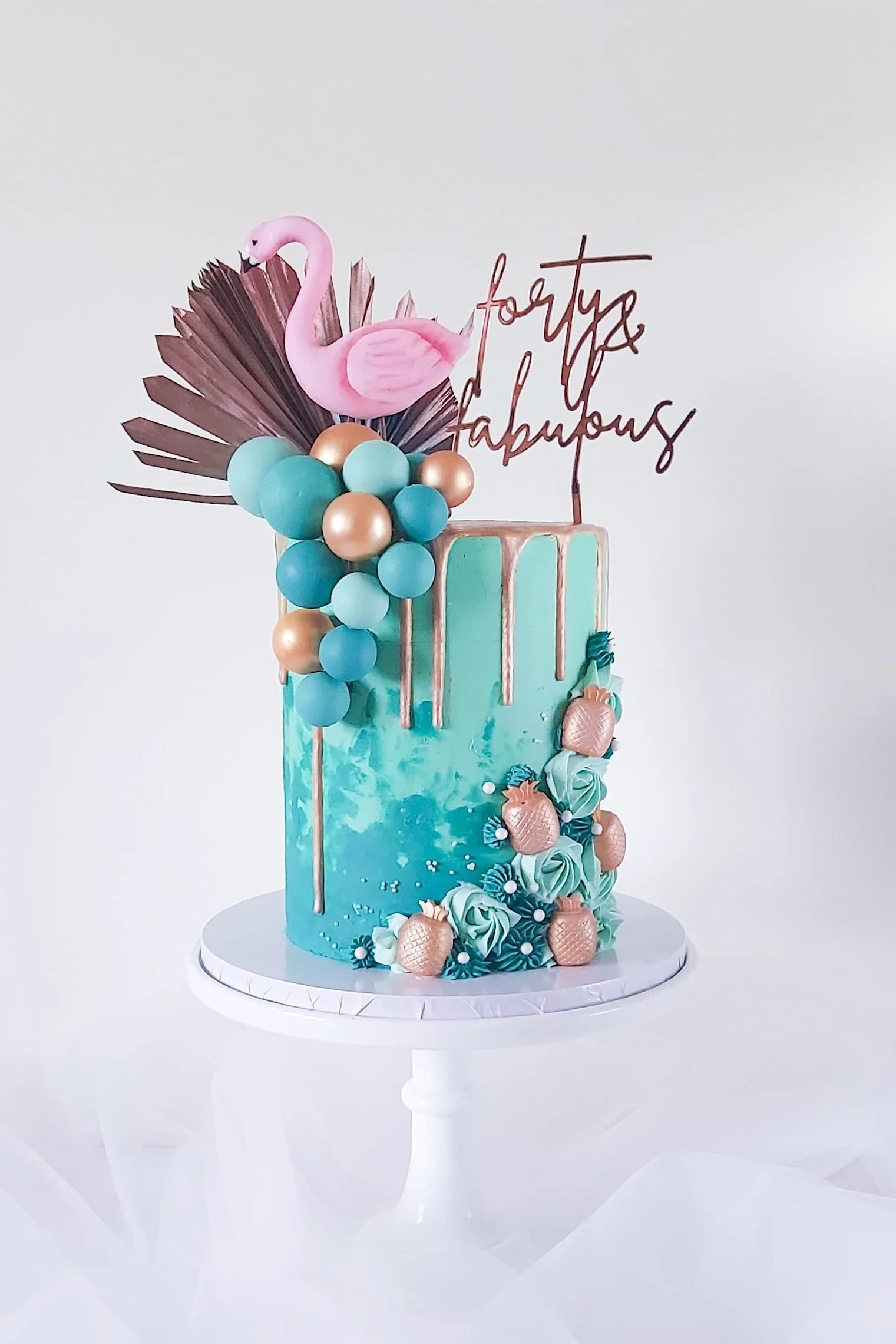 A tall, turquoise birthday cake with pink and gold accents, decorated with a pink flamingo topper, balloons, gold pineapples, and a pink 'birthday fabulous' sign, on a white cake stand.