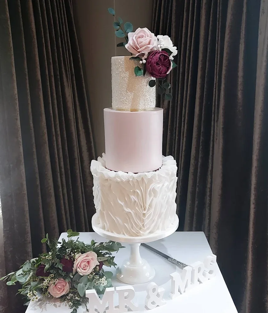 A three-tier wedding cake with a flower arrangement on top, surrounded by a bouquet and Mr. & Mrs. signs, set in front of dark curtains.