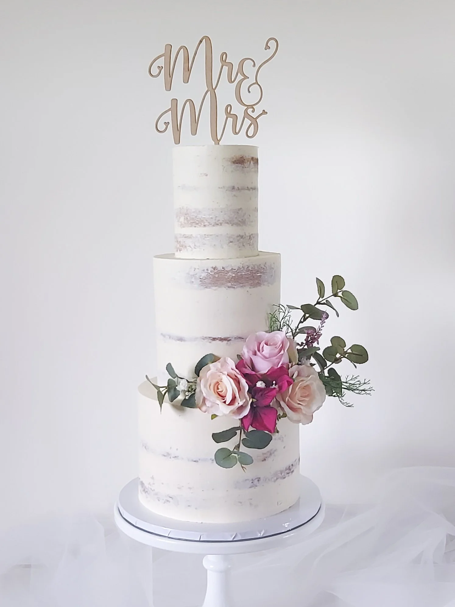 A three-tier wedding cake with semi-naked white frosting, decorated with pink and white roses, green foliage, and a 'Mr & Mrs' cake topper.
