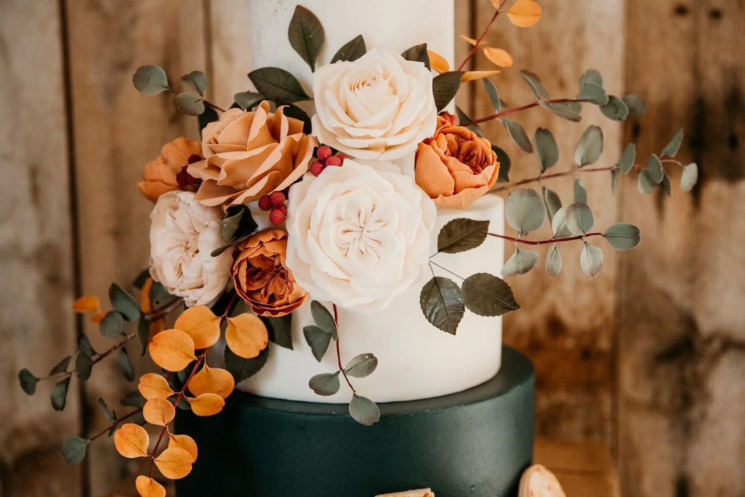 A decorative cake adorned with a floral arrangement of white and orange roses, green leaves, and red berries, set against a rustic wooden background.