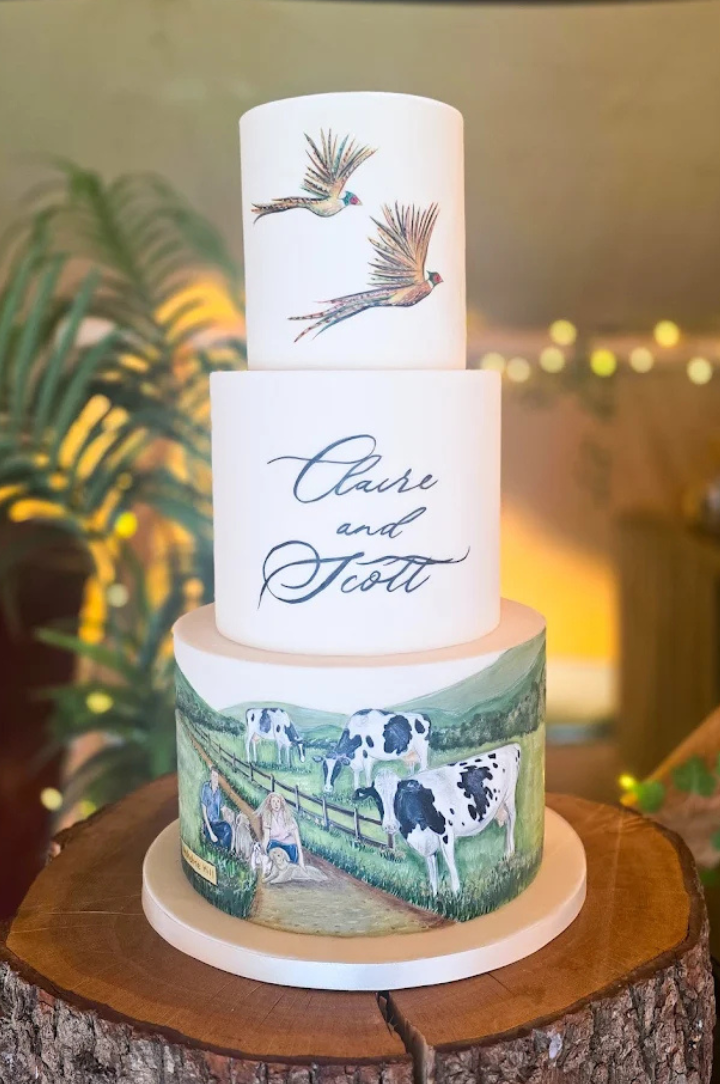 Three-tiered wedding cake with hand-painted cows, a barnyard scene, two flying birds on the top tier, and the names Claire and Scott written on the middle tier. The cake sits on a wooden slab.