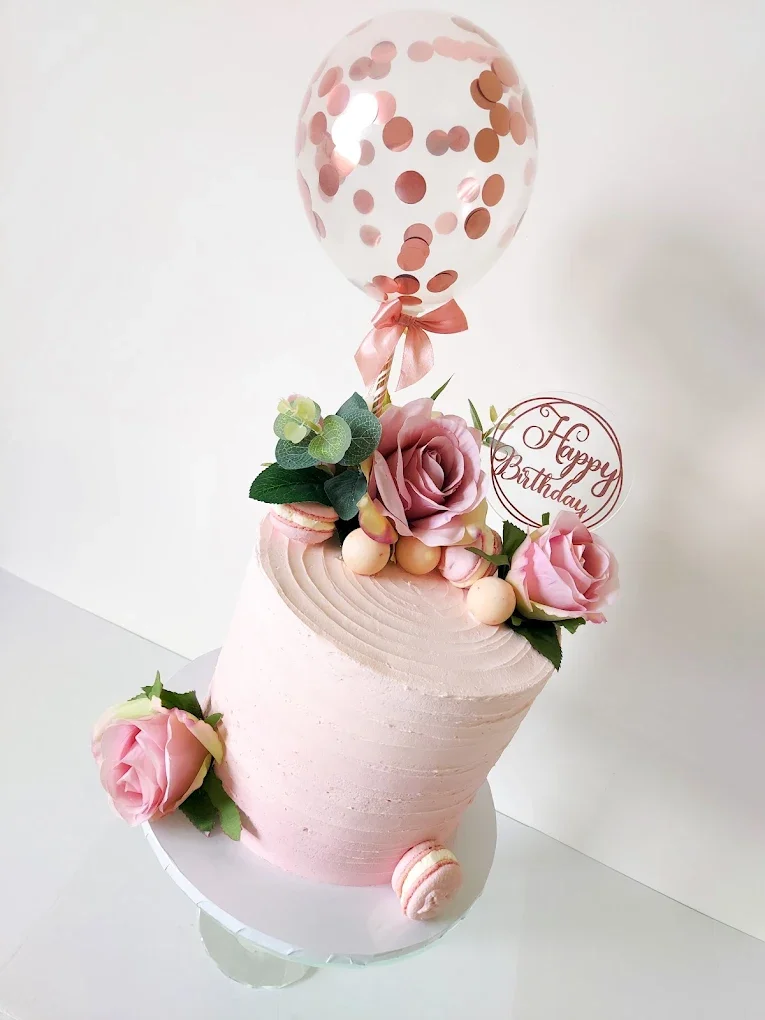 A pink birthday cake decorated with roses, green leaves, macarons, and a transparent balloon with pink confetti, with a round 