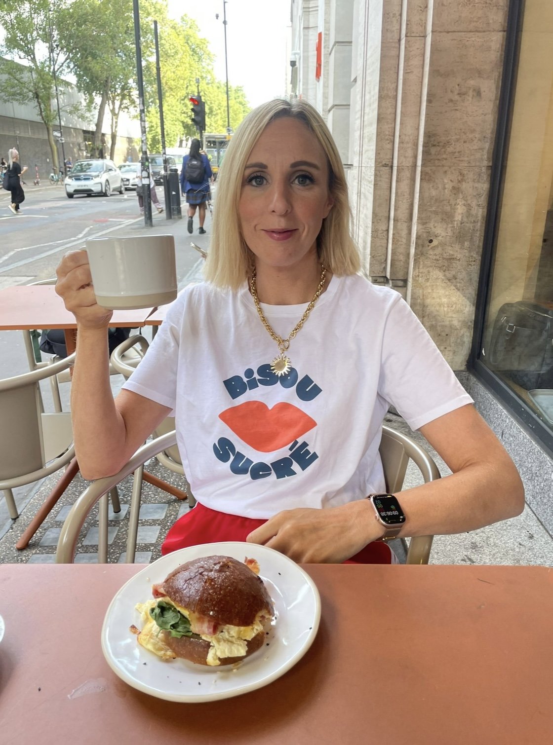 A woman sitting at an outdoor cafe with a city street in the background. She is holding a large white mug. She is wearing a white t-shirt with orange lips and blue lettering, a gold necklace, and a smartwatch. There is a sandwich with eggs, bacon, lettuce, and tomato on a white plate in front of her.