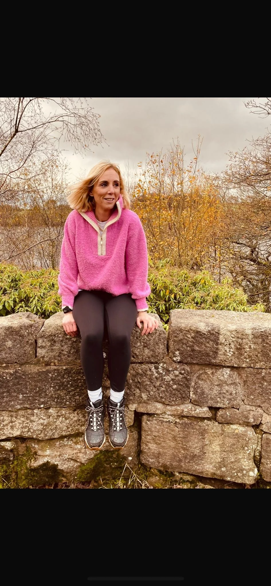A woman sitting on a stone wall outdoors, wearing a pink fleece pullover, black leggings, hiking boots, and white socks, with trees and shrubs in the background on a cloudy day.