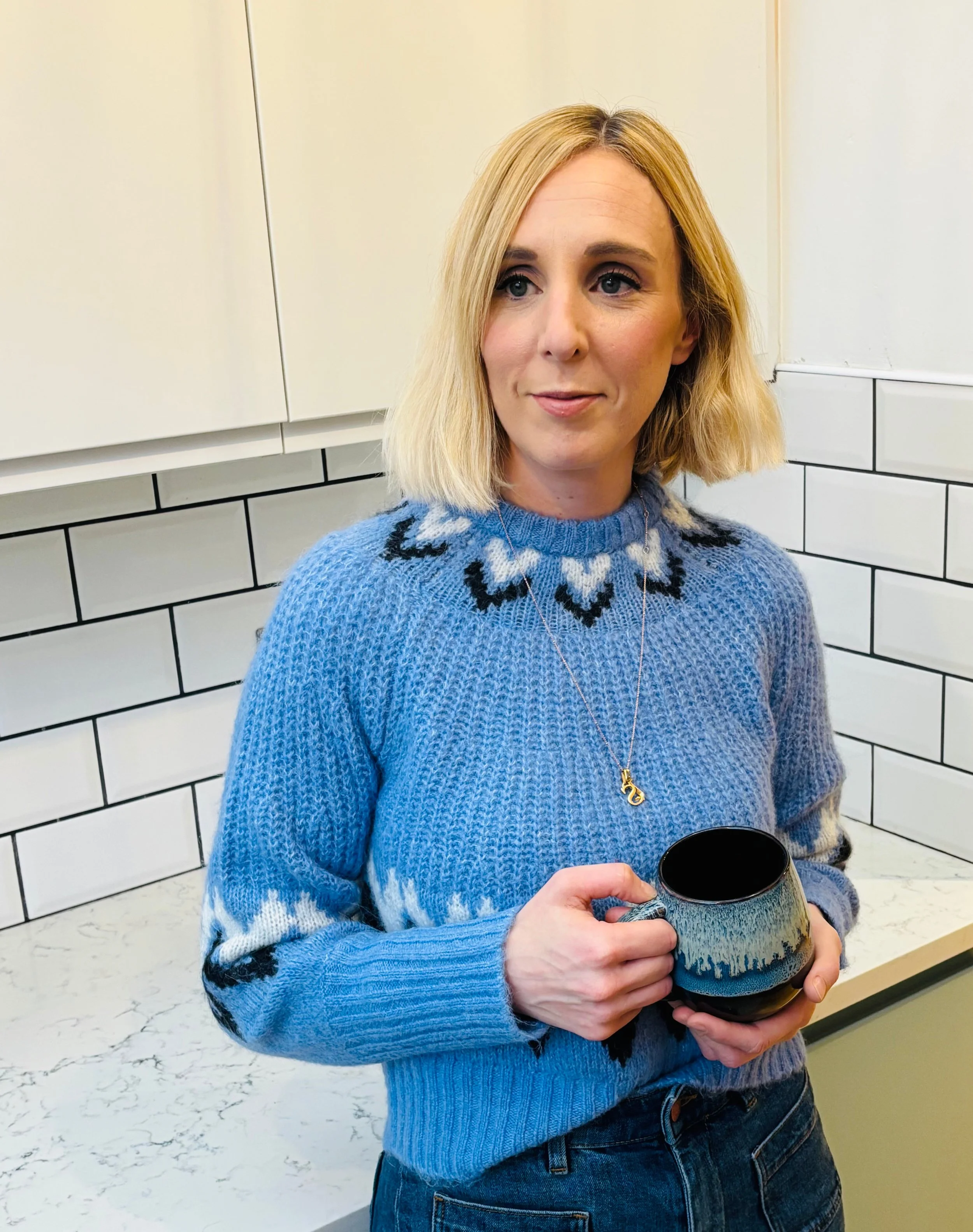 A woman with blonde hair wearing a blue sweater with a black and white pattern, holding a ceramic mug, standing in a kitchen with white tiled walls.