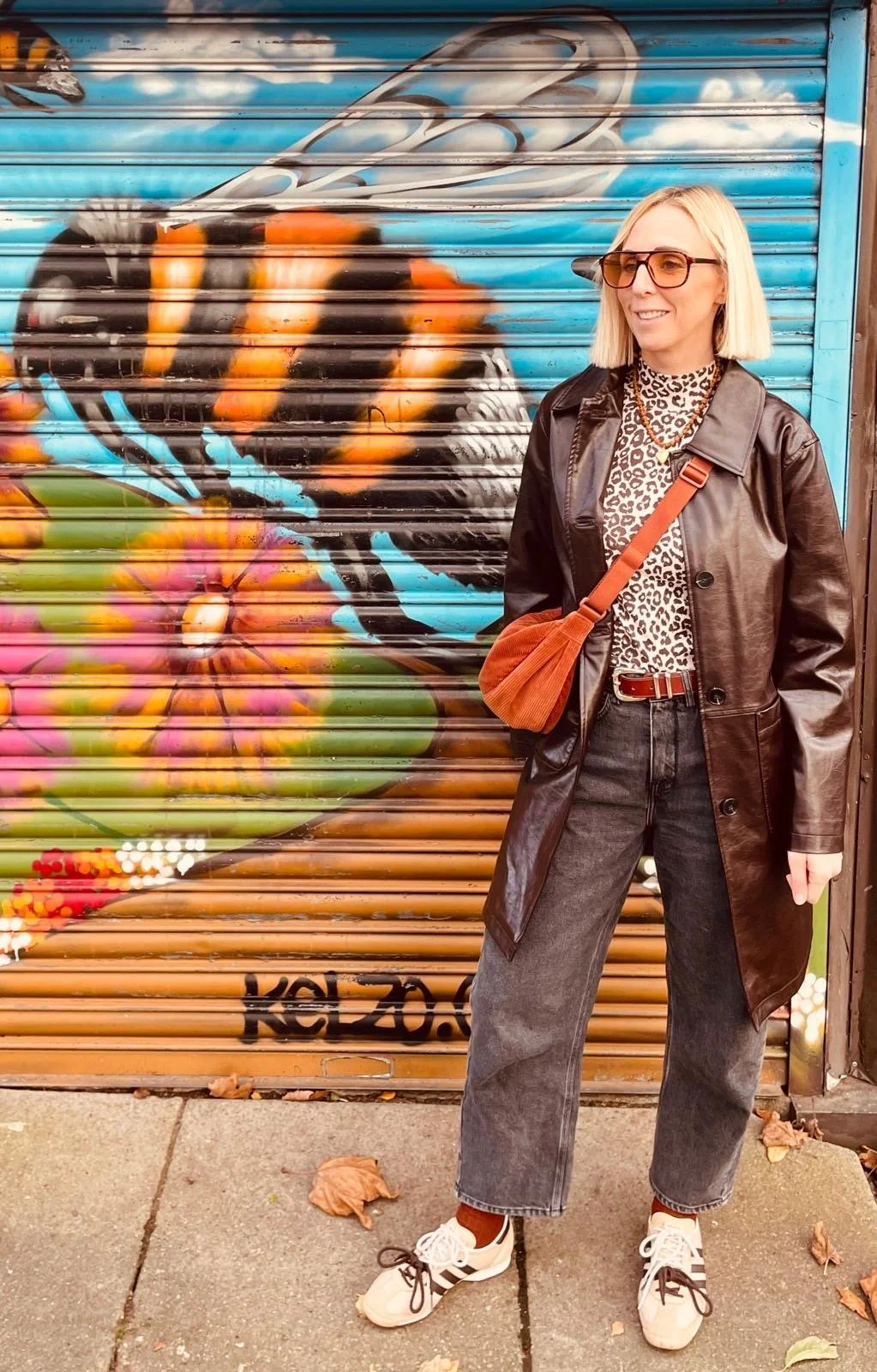 A woman with blonde hair, glasses, and a leopard print top, wearing a black leather coat, gray jeans, and sneakers standing in front of a colorful graffiti mural of a butterfly and a flower on a metal shutter.