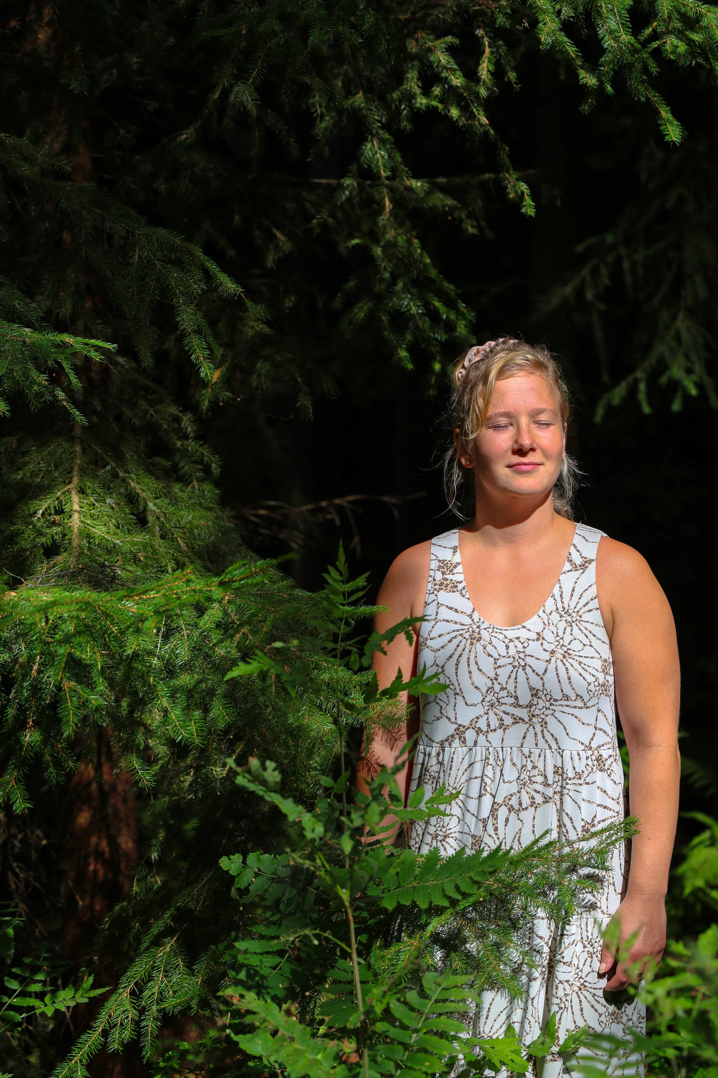 Nuori nainen seisoo metsän keskellä auringonvalossa, ympärillään vihreitä kasveja ja puita.