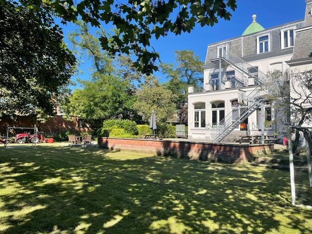 Luxe witte villa met groene koepel en buitentrap, omgeven door groene bomen en een tuin met schaduw.