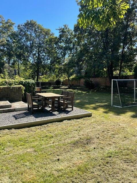 Een tuin met een houten tafel en stoelen op een betegeld terras, een grasveld ernaast, omgeven door bomen en een hek. In de rechterhoek een voetbalgoal.