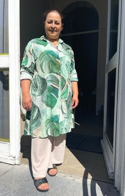 Vrouw staat buiten bij de toegangsdeur, draagt een groene, blauwe en witte jurk en witte broek, smiling.