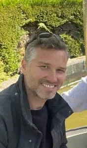 Een man met kort haar en een baard, lachend, buiten met groene planten op de achtergrond.