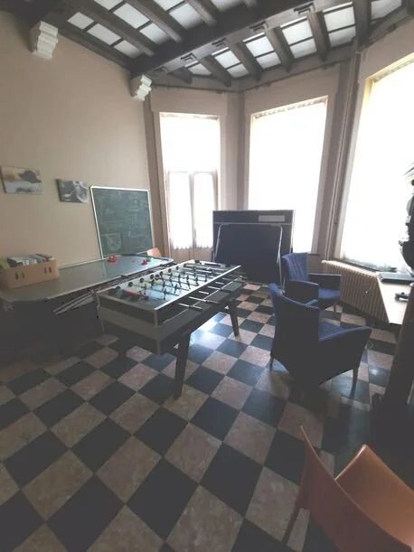 Kamer met grote ramen, een tafel met een spelcomputer, twee blauwe stoelen en een schoolbord.