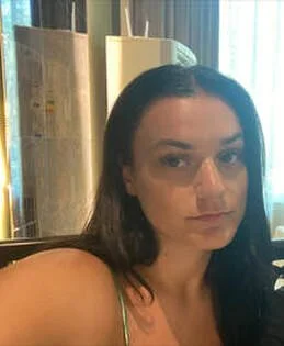 Close-up van een vrouw met donker haar, zittend in een interieur met gordijnen en houten decoratie.