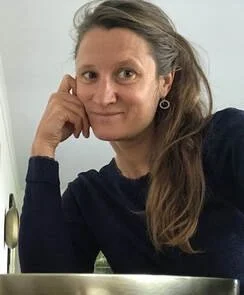 Een vrouw met bruin haar en een navyblauwe trui, zittend aan een tafel op een lichte achtergrond.