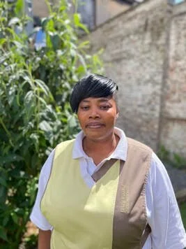 Vrouw met kort zwart haar, witte blouse, lichtgele en beige vest, buiten met groen en een muur op de achtergrond.