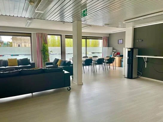 Binnenruimte met banken, een eettafel met stoelen, een waterkoeler en een koelkast, grote ramen met uitzicht op bomen buiten.