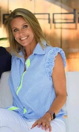 Vrouw met blond haar, blauwe gestreepte blouse met ruffles, lachend, zittend in een lichte ruimte met ramen.