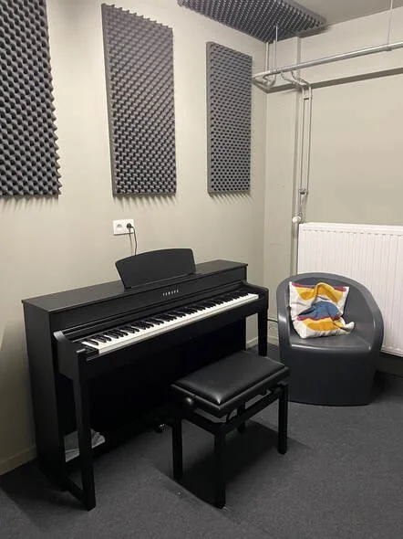 Een zwart digitale piano met bijbehorend bankje, een comfortabele stoel met een kussen, en geluidsabsorberende panelen aan de muur in een muziekstudio.