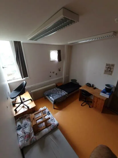 Kamer met bed, bureau, stoel en dekbed op vloer, kleine raampje, en lampen aan plafond.