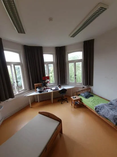 Een slaapzaal met één bed met groen dekbed en één bed zonder beddengoed, een bureau met een stoel en kleine voorwerpen, grote ramen met donkere gordijnen, houten vloer, witte muren en plafond met verlichtingsbuizen.