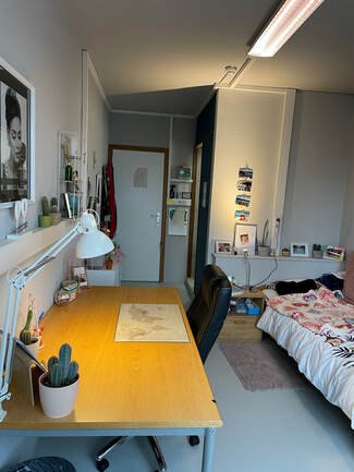 Een slaapkamer met een bureau, stoel, bed, en diverse decoraties en boeken.