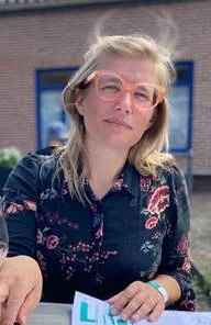 Een vrouw met blond haar, bril en een donkere bloemenprint blouse, zittend buiten.