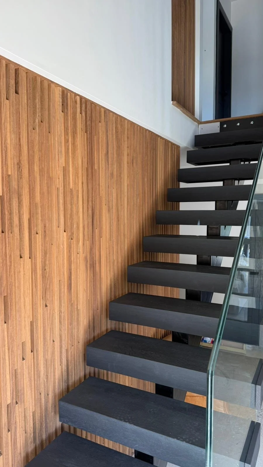 Interior view of a modern staircase with black floating steps, a glass railing, a wood-paneled wall, and a white wall with a wooden accent in the background.