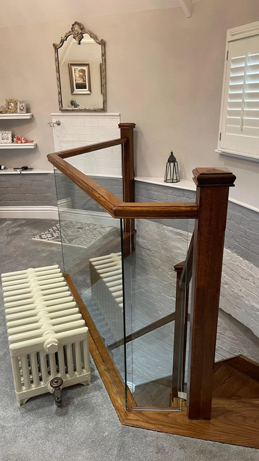 Corner view of a staircase with glass railing and wooden handrail, white-painted brick walls, a window with white shutters, and decorative items on shelves and ledge.