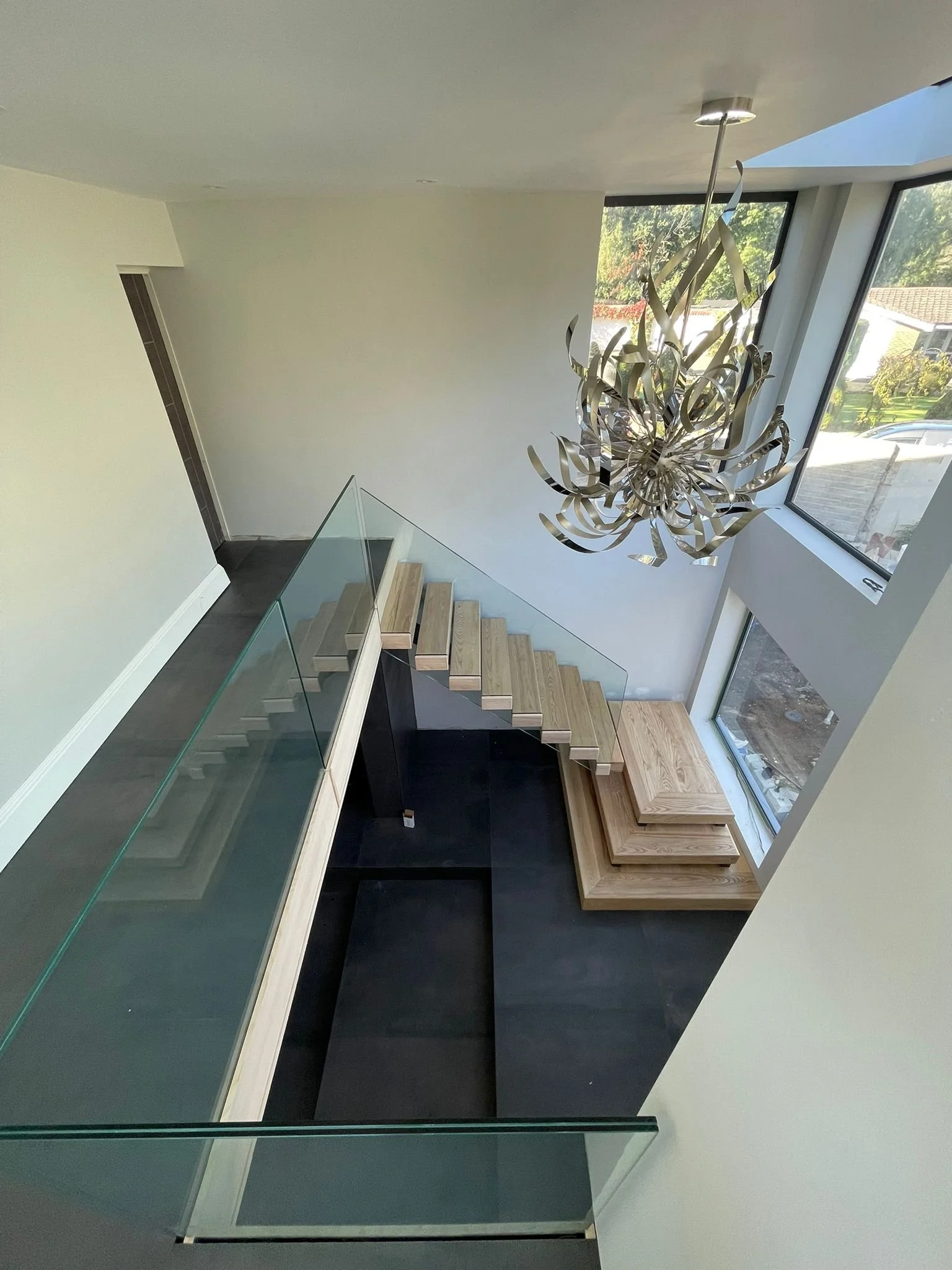 Interior view of a modern staircase with glass railings, wooden steps, large windows letting in natural light, and a decorative chandelier hanging from the ceiling.