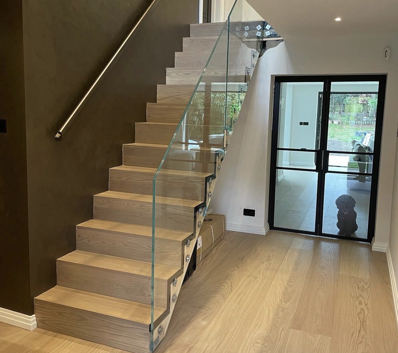 Interior view of a modern staircase with wooden steps, glass railing, and a sliding glass door leading to a backyard.