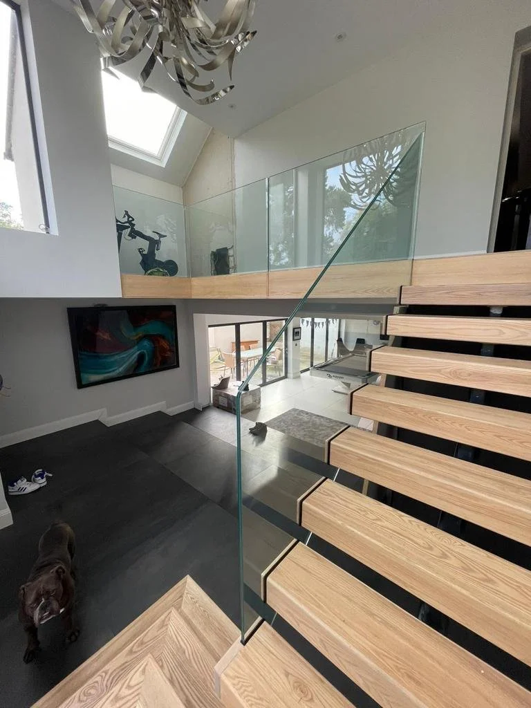 Interior view of a modern house showing a staircase with wooden steps and a glass railing, a dog on the dark floor, a wall-mounted TV, and large windows with a view outside. There's also a second-floor loft with a bike and a chandelier hanging from t