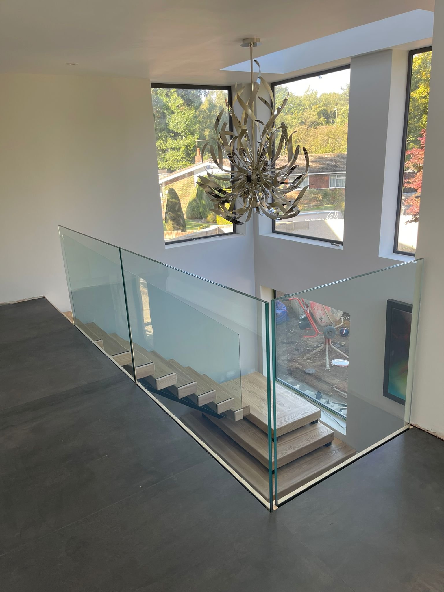Interior view of a modern home with a staircase enclosed by glass railings, large windows, and a contemporary chandelier hanging from the ceiling.