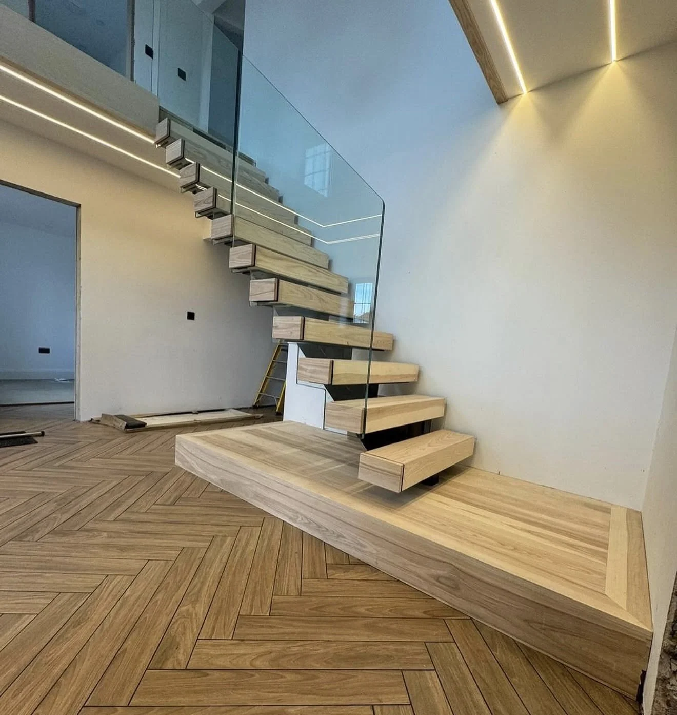 Unfinished modern staircase with floating wooden steps, glass railing, and herringbone wooden floor in a contemporary interior