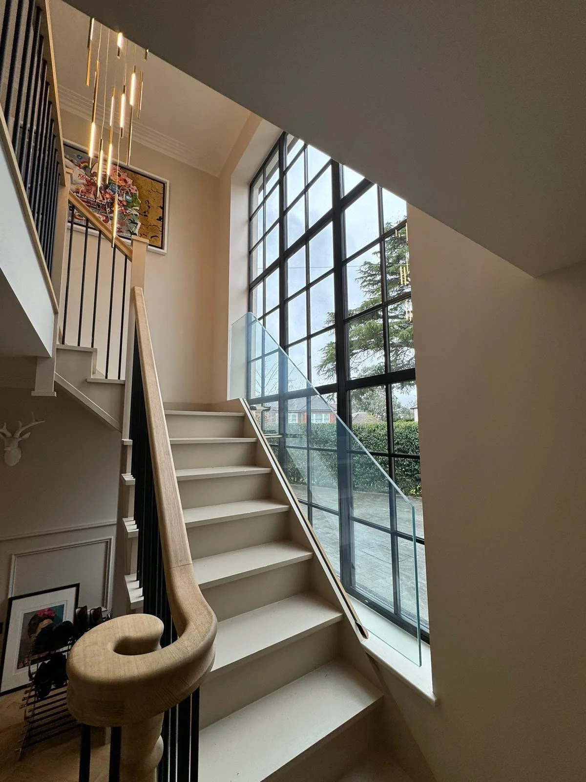 Modern staircase with light wood handrail, black metal balusters, and large floor-to-ceiling glass window showing an outdoor view with trees and a cloudy sky.