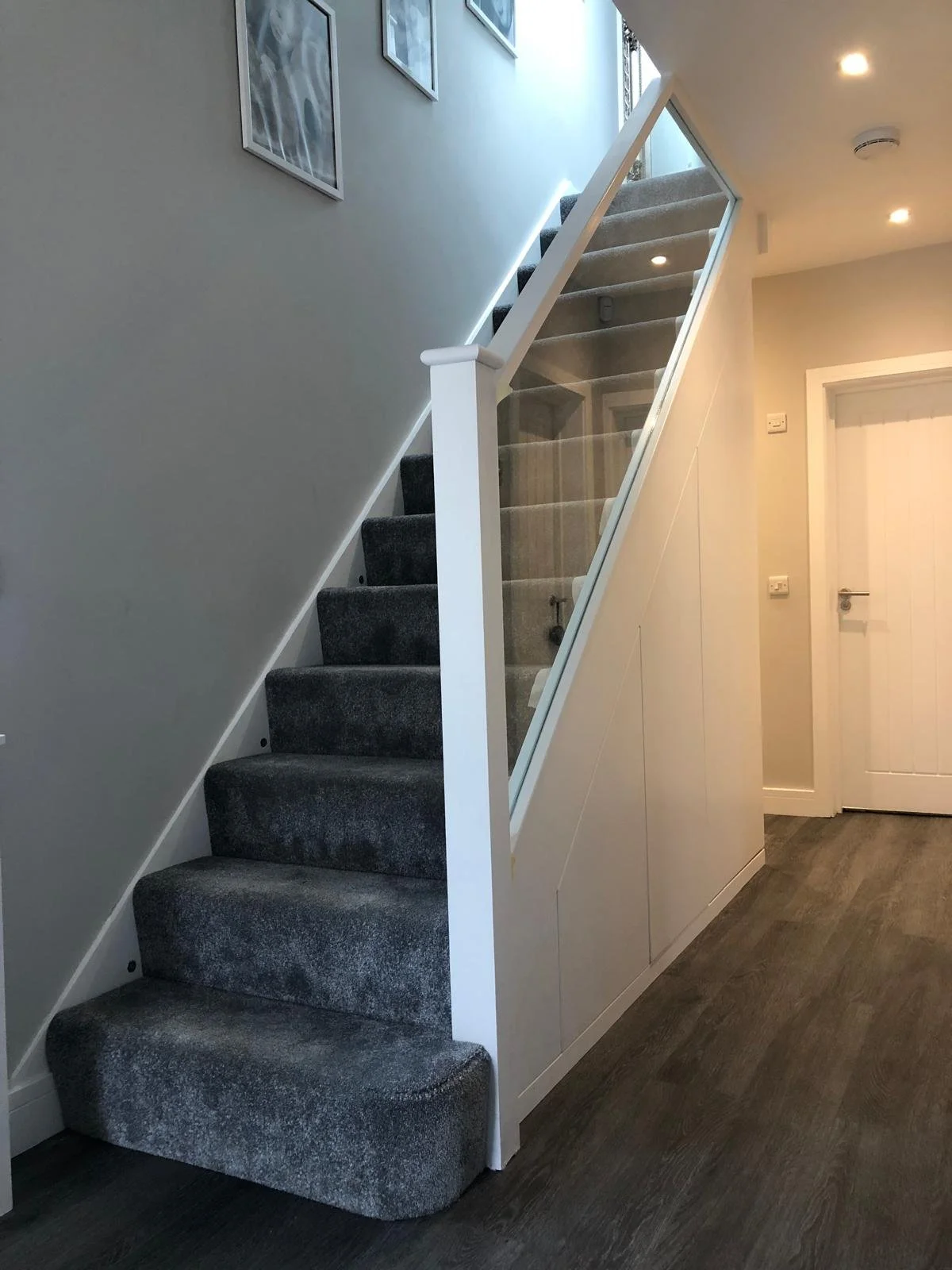 Interior staircase with a partial glass railing leading up, dark carpeted steps, and a white wall with framed pictures. The floor is dark wood, and there is a white door at the bottom.