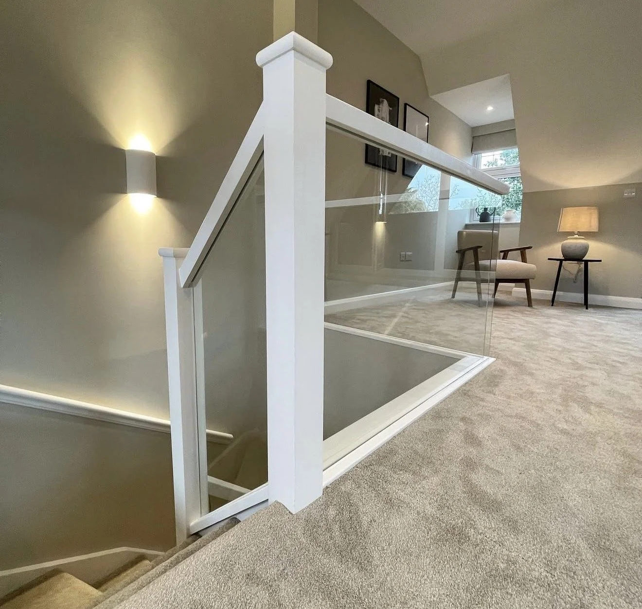 Interior of a modern home with a staircase, glass railing, beige carpet, and a sitting area with a chair, side table, and lamp.