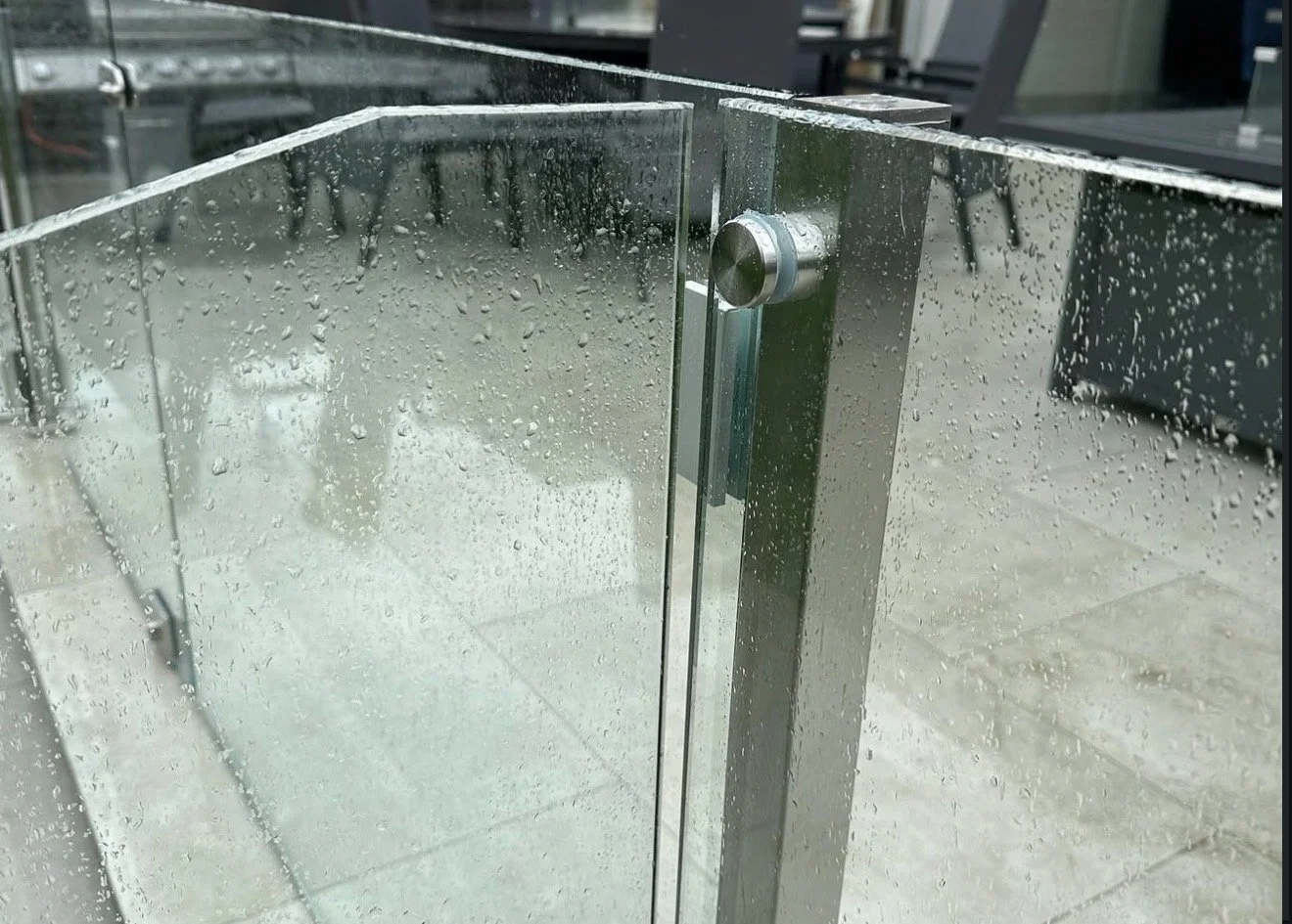 Close-up of a glass shower partition with water droplets and metal hardware, with a blurred background.