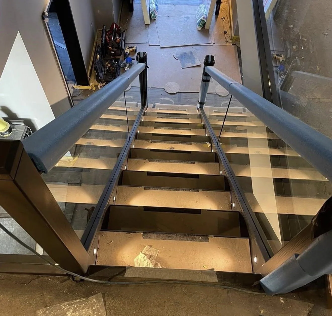 View from the top of a staircase looking down, with glass sides and metal railings, leading to a construction area with tools and materials on the floor.