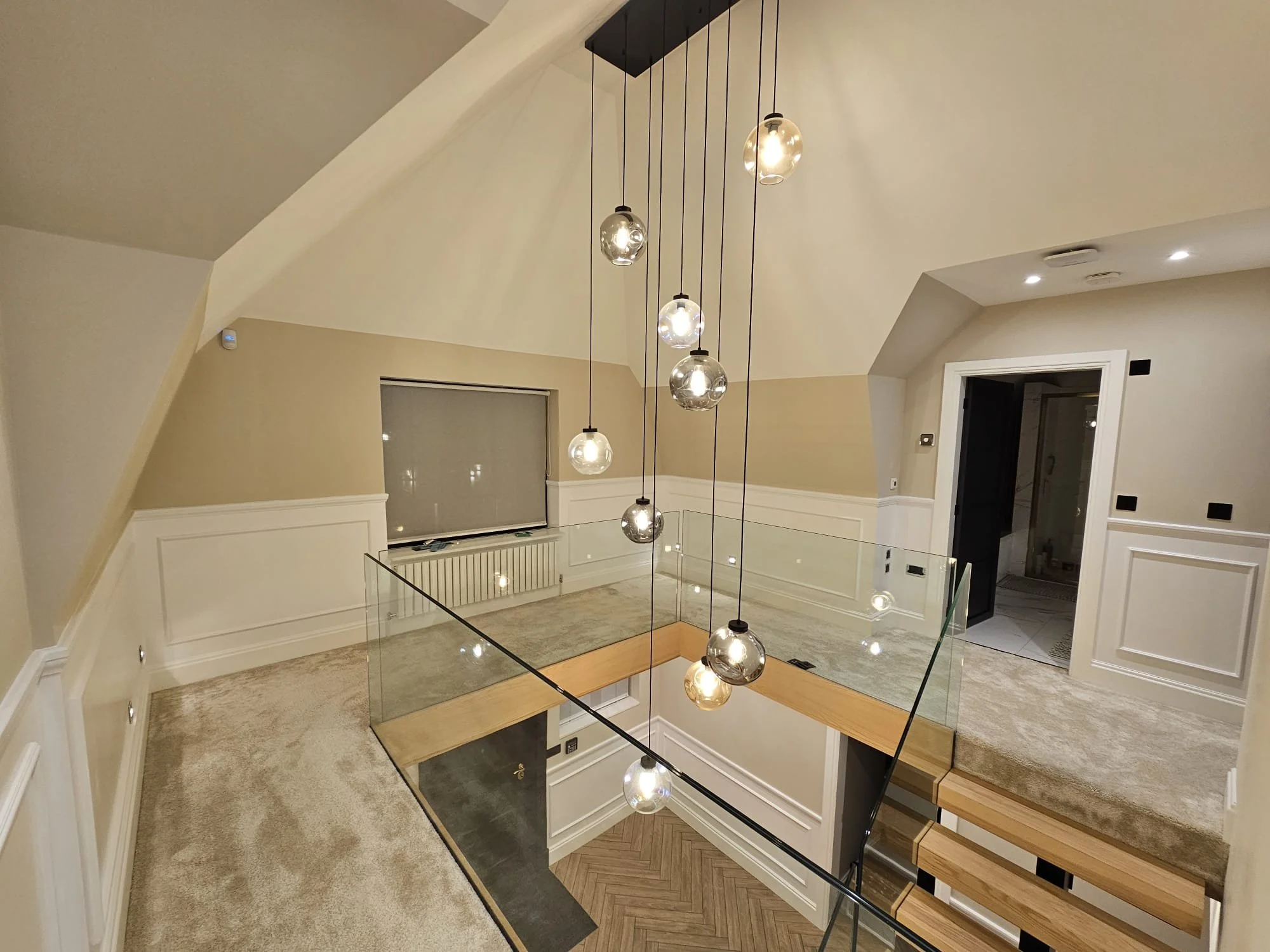 Interior view of a modern home staircase area with hanging spherical pendant lights, beige walls, white wainscoting, carpeting, glass railing, and a staircase leading down to a lower floor.