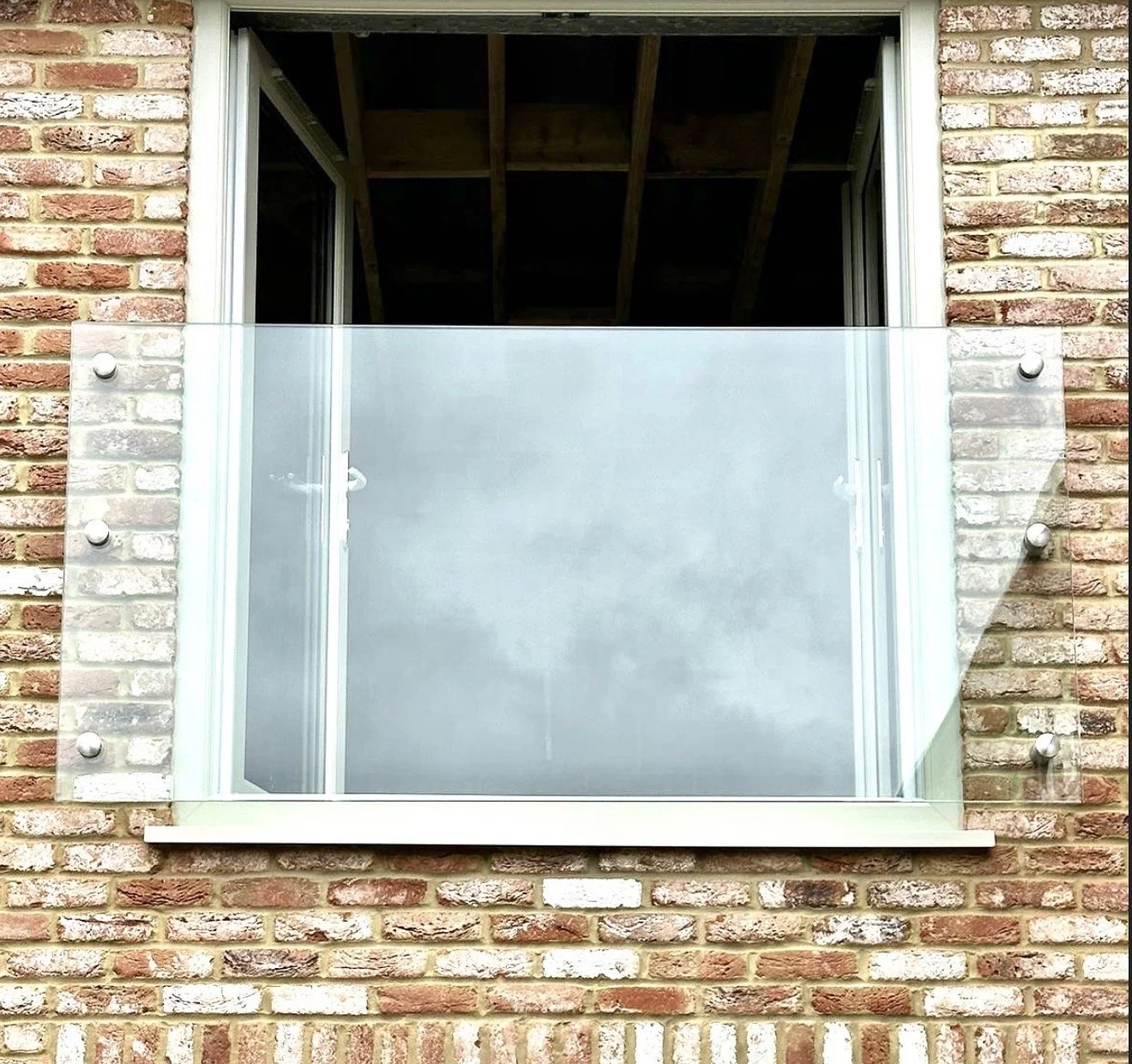 A glass balcony railing installed on a brick building, with a window above and construction wood visible inside the building.