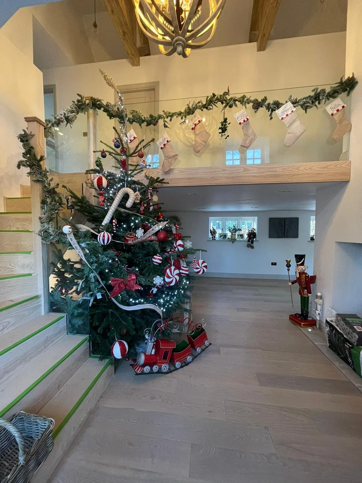 A decorated Christmas tree with red, white, and green ornaments, candy canes, and a red bow, situated next to a staircase in a living room. Above the tree, a garland with stockings, each personalized with a name, is hung across the railing. There are