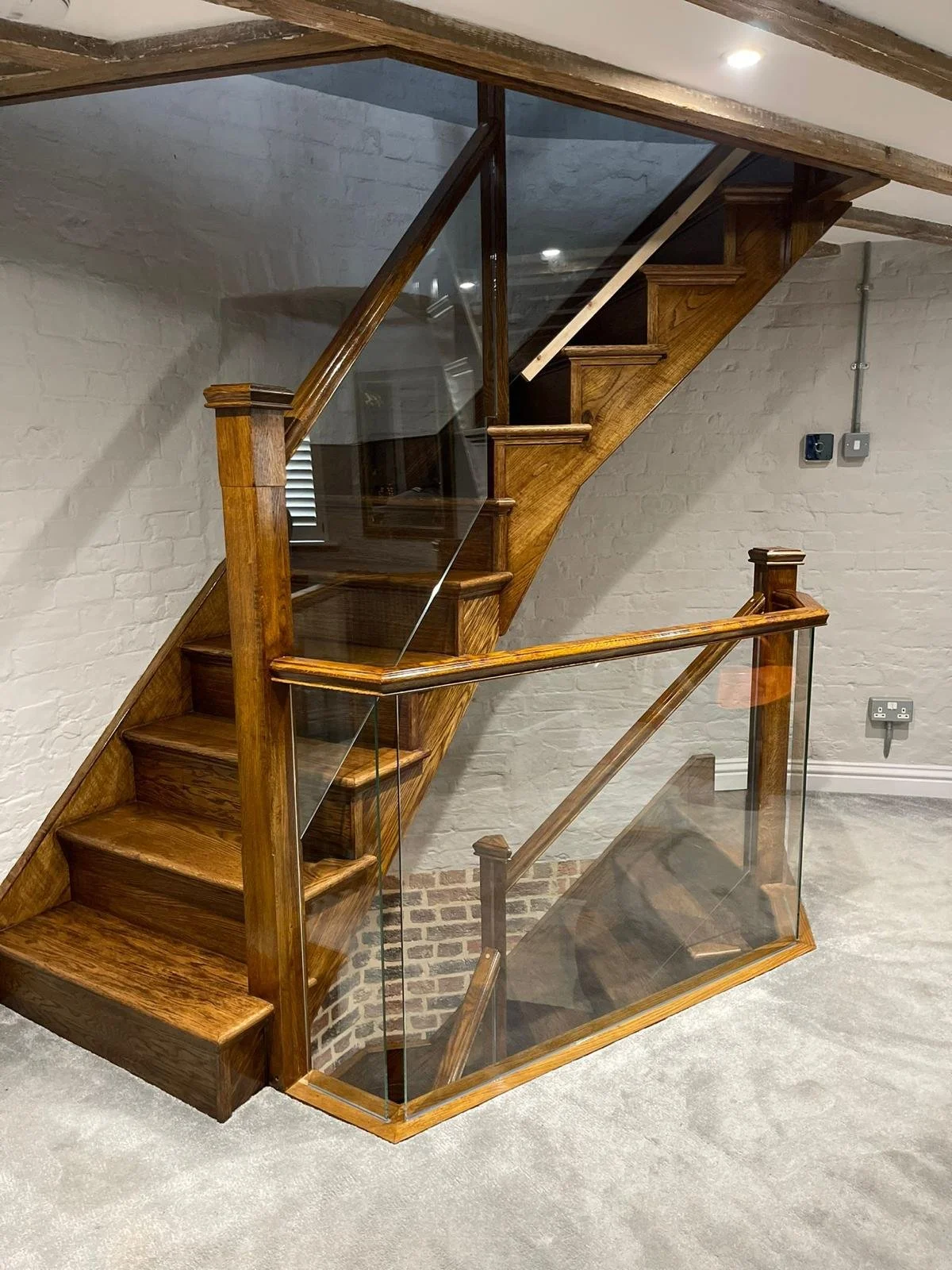 Wooden staircase with glass railing in a basement with white painted brick wall and gray carpet