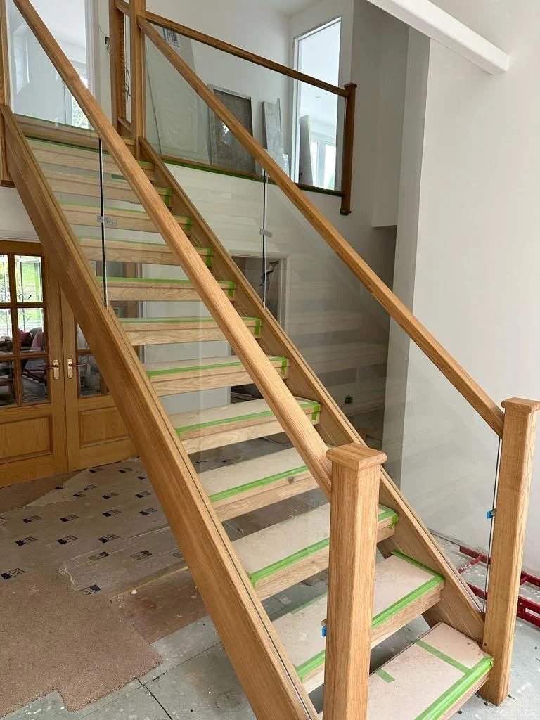 Wooden staircase under construction with protective green tape on steps, glass panels on sides, in a modern home interior, with a window and double doors in the background.