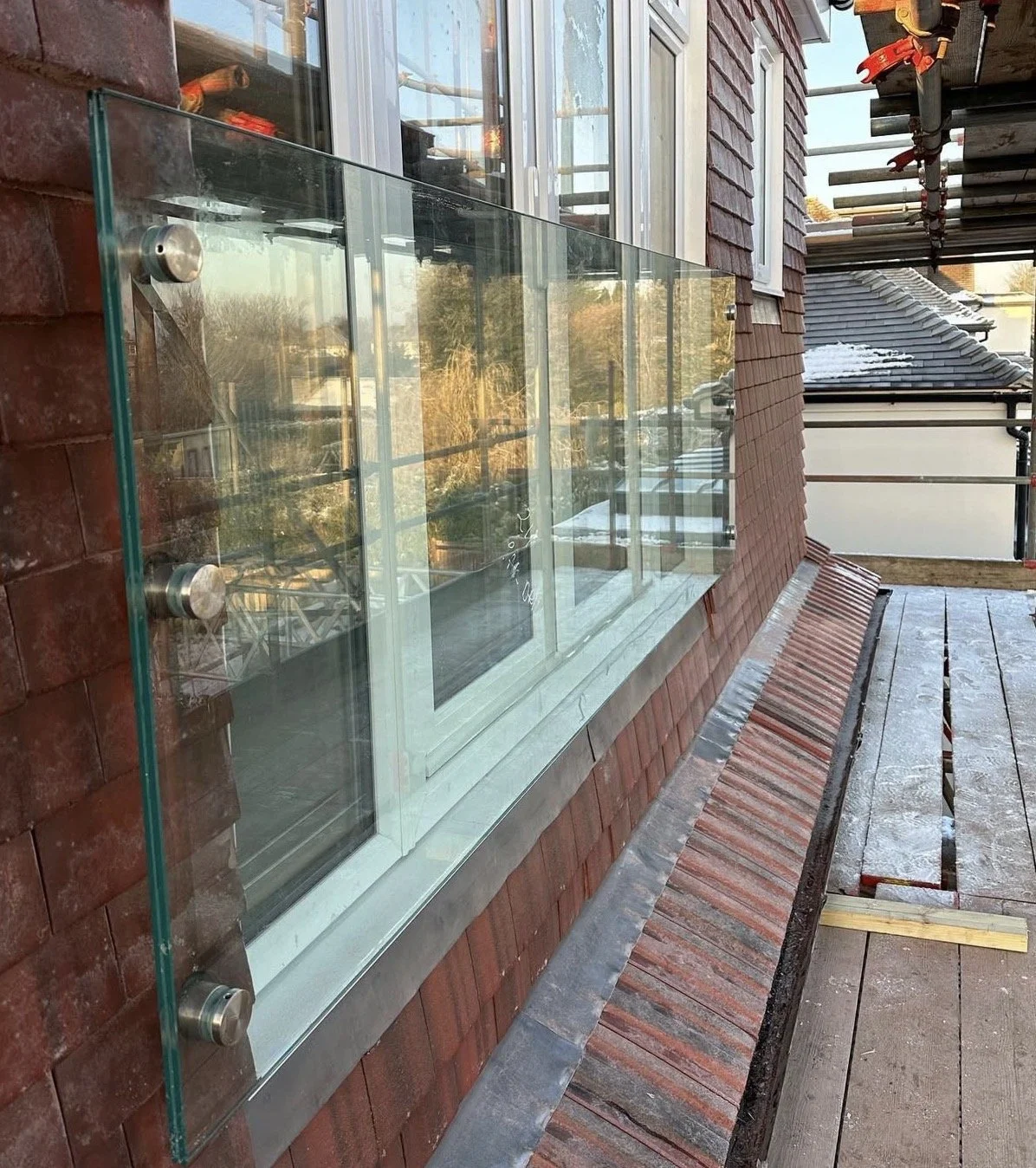 View of a building under construction with a glass railing on a balcony or ledge, brick exterior, and a floor with wooden planks, adjacent to windows and scaffolding.