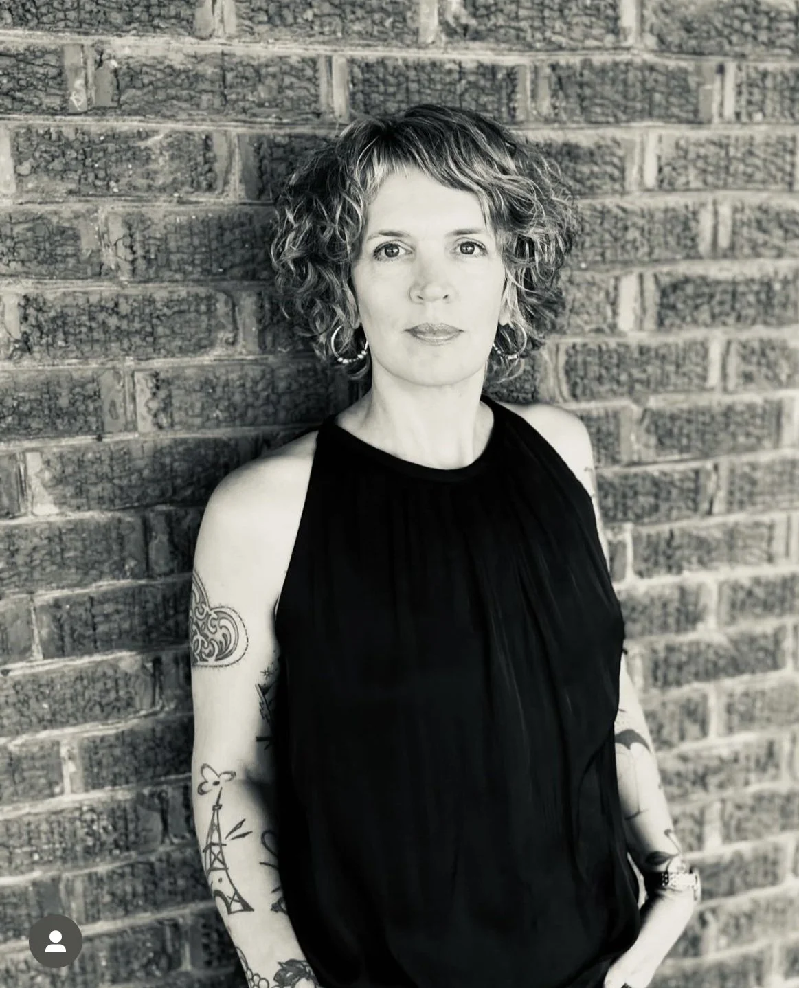 Black and white photo of a woman with curly hair, wearing a sleeveless black top, standing against a brick wall, with tattoos on her arms.