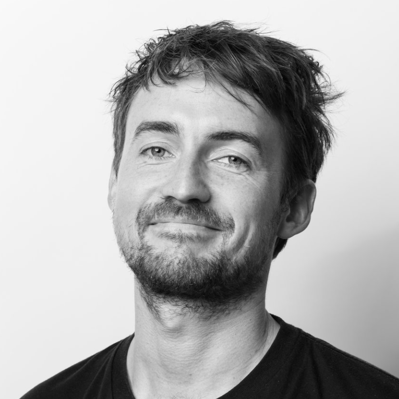 Sort-hvid portrætfotografi af en mand med kort, rodet hår, skæg og et smil, iført sort t-shirt, mod en lys baggrund.